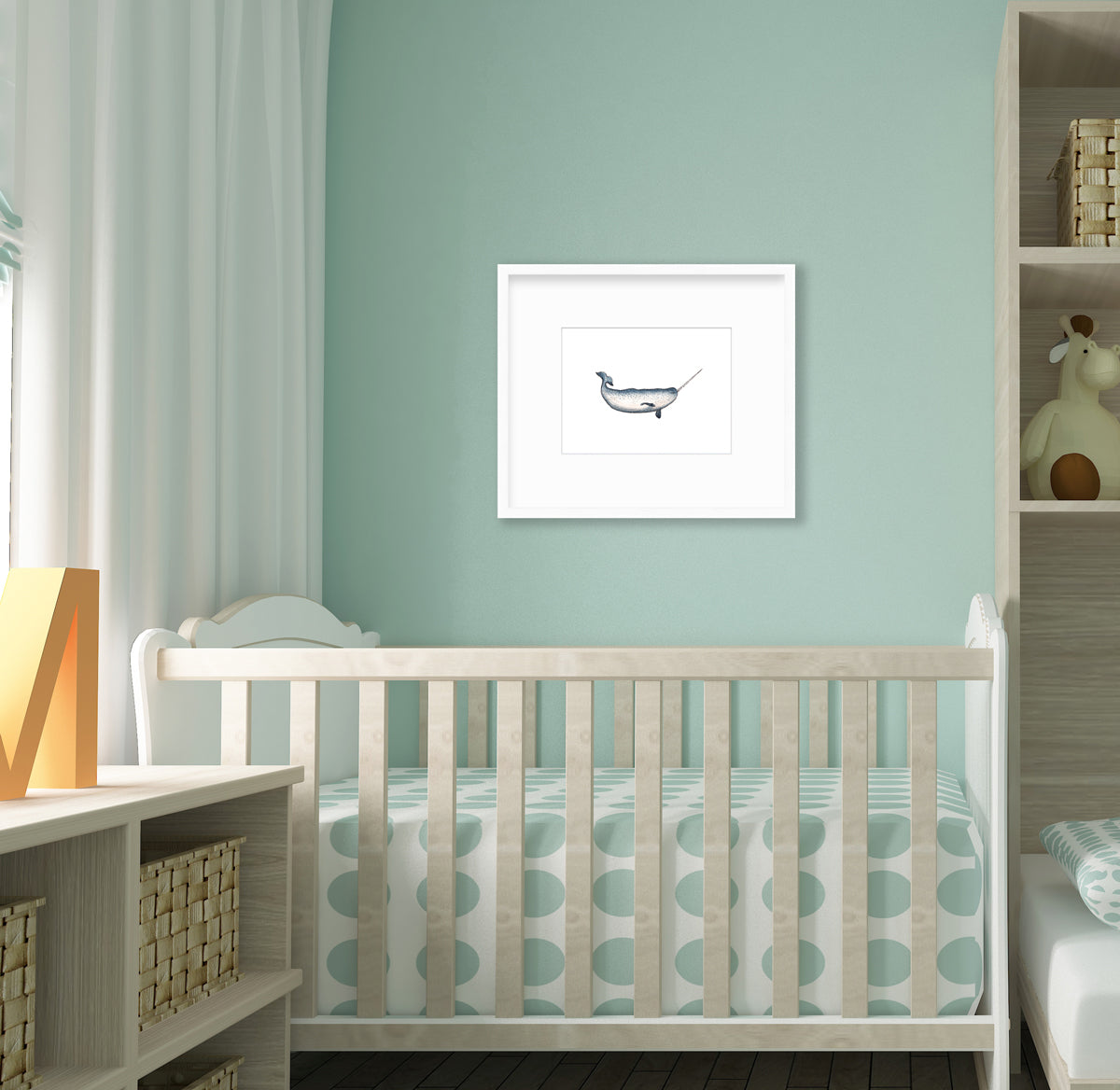 A blue and white narwhal print in a white frame with a mat hangs on a sea foam green wall above a natural wood crib with light green dotted bedding and next to a tall bookshelf with decorative accessories.