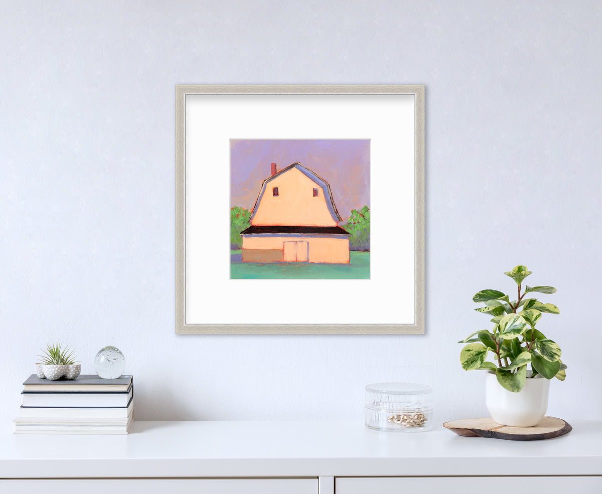 A print of a light orange barn, a lavender sky, green bushes and a teal ground in a silver frame with a mat hangs on a white wall above a white surface with a stack of books, a clear round container with gold accessories and a potted plant.