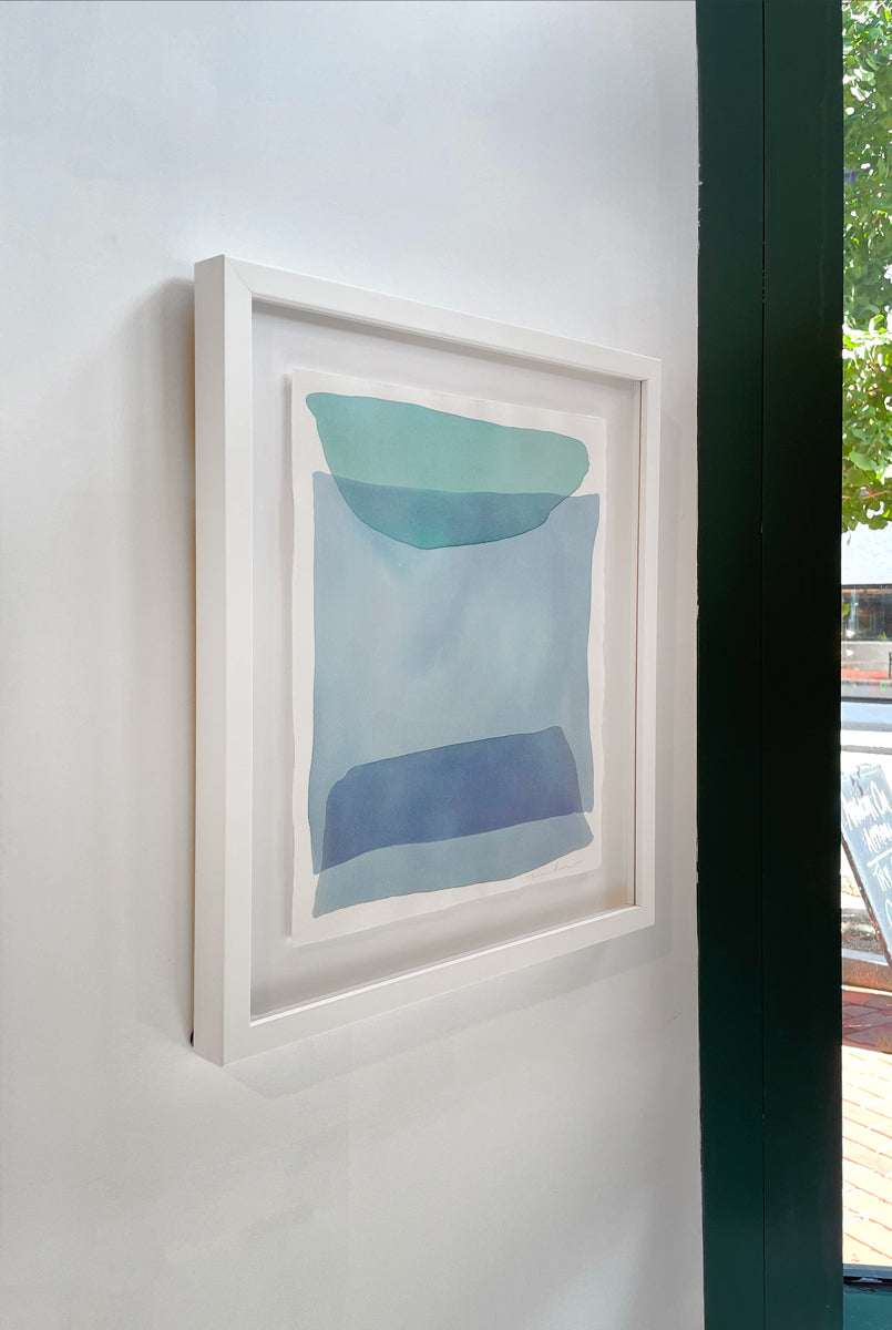 A left side-angle view of blue and green shapes overlap in this painting by Nealy Hauschildt in a white frame floating above a white matte hangs on a white wall.