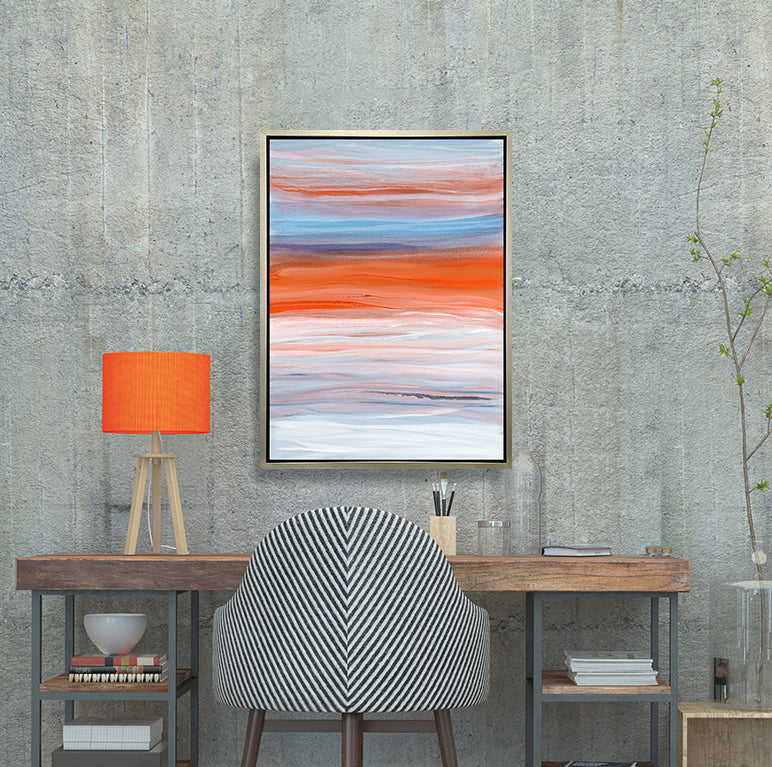 A blue, orange, light pink and white abstract print in a silver floater frame hangs on a concrete wall above a wood desk with a lamp with an orange shade, stacks of books, a large glass bottle with a greenery stem.