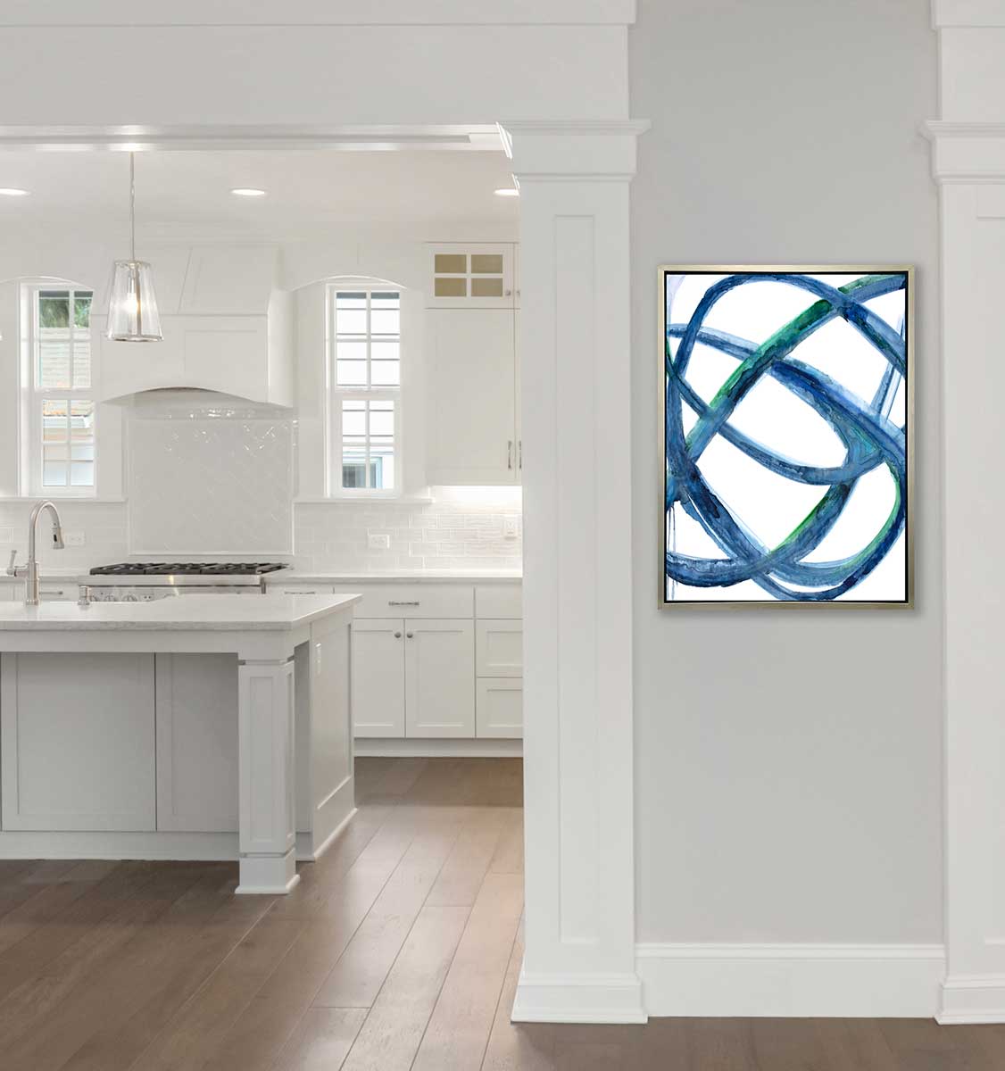 A blue, green and white abstract print in a silver floater frame hangs on a light grey wall next to an opening to a white kitchen.