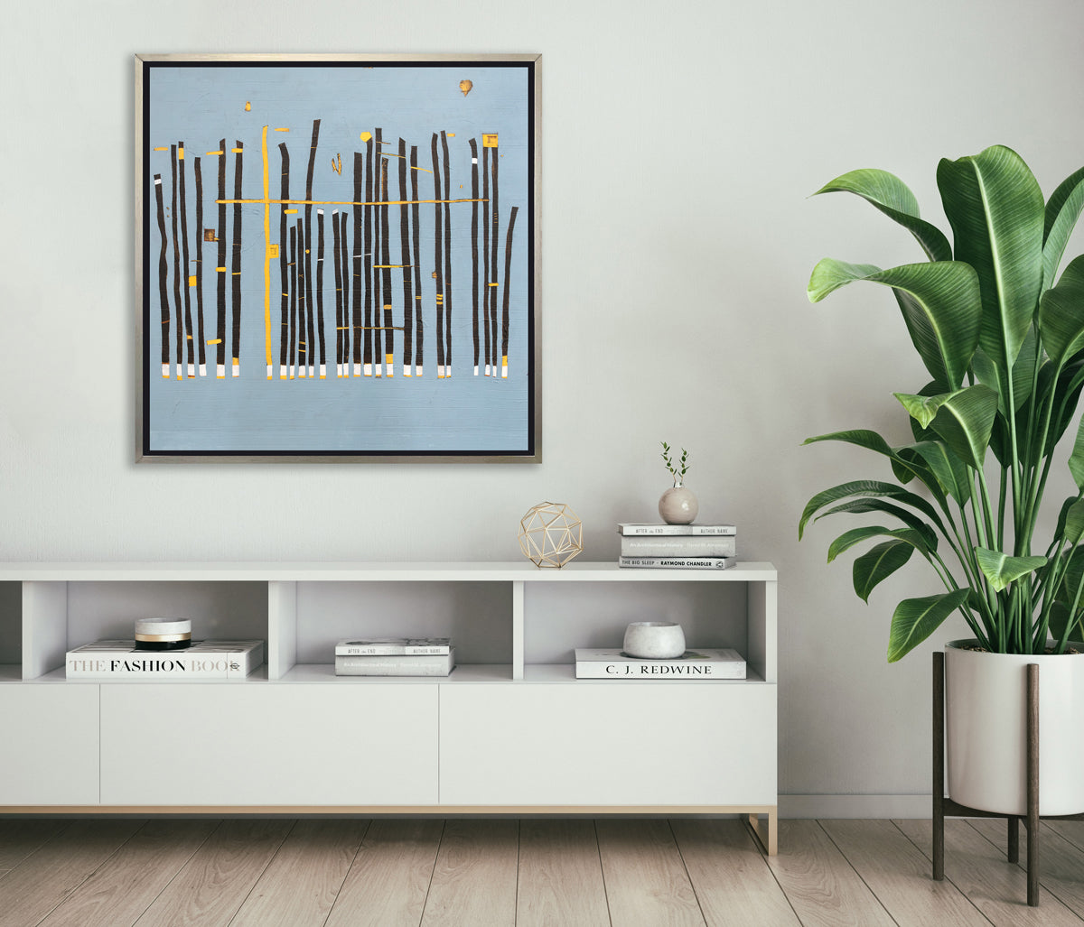 A blue, black and yellow abstract print in a silver floater frame hangs on a white wall above a white console cabinet with a gold wire sphere, a stack of books and a tall potted floor plant.