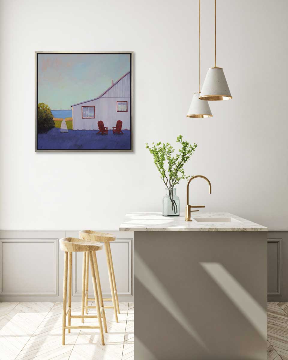 A blue, lavender and light purple contemporary seascape print with a beach house, two red adirondack chairs and a walkway leading to the water in a silver floater frame hangs on a white wall above a white marble topped kitchen island with a sink, a clear vase with green stems and two wood counter stools.