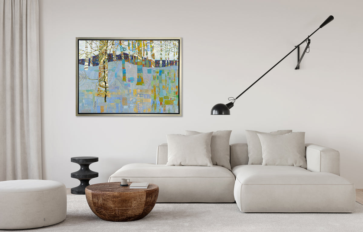 A multicolored abstract landscape in a silver floater frame hangs on a white wall above a white sectional sofa with pillows, a black wall mounted adjustable lamp, a rounded wood coffee table, a black pedestal side table and a large white rounded ottoman.