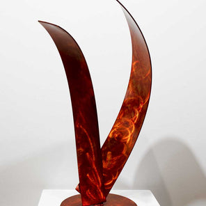 The left side of abstract, steel sculpture with orange dye forming a shape of a wave, sitting on a pedestal in front of a white wall.