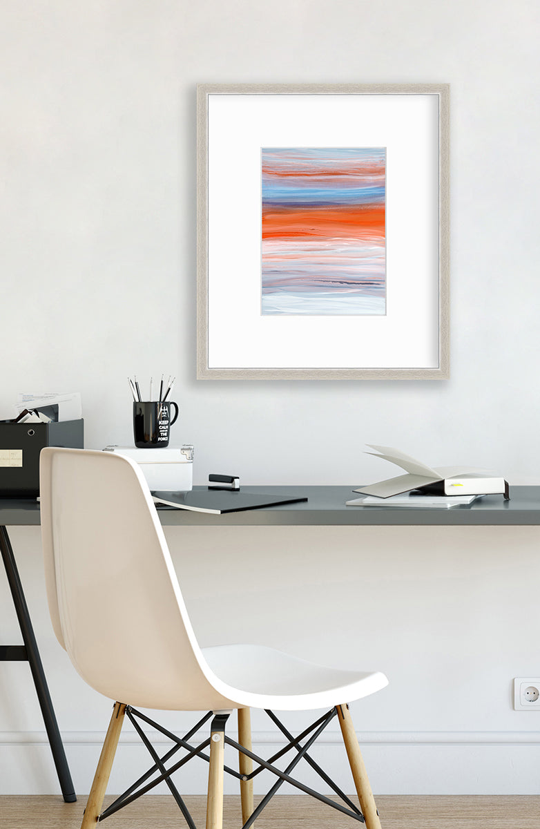 A blue, orange, light pink and white abstract print in a silver frame with a mat hangs on a white wall above a grey desk with an open book, a few small boxes, a black mug with pencils and a white chair.