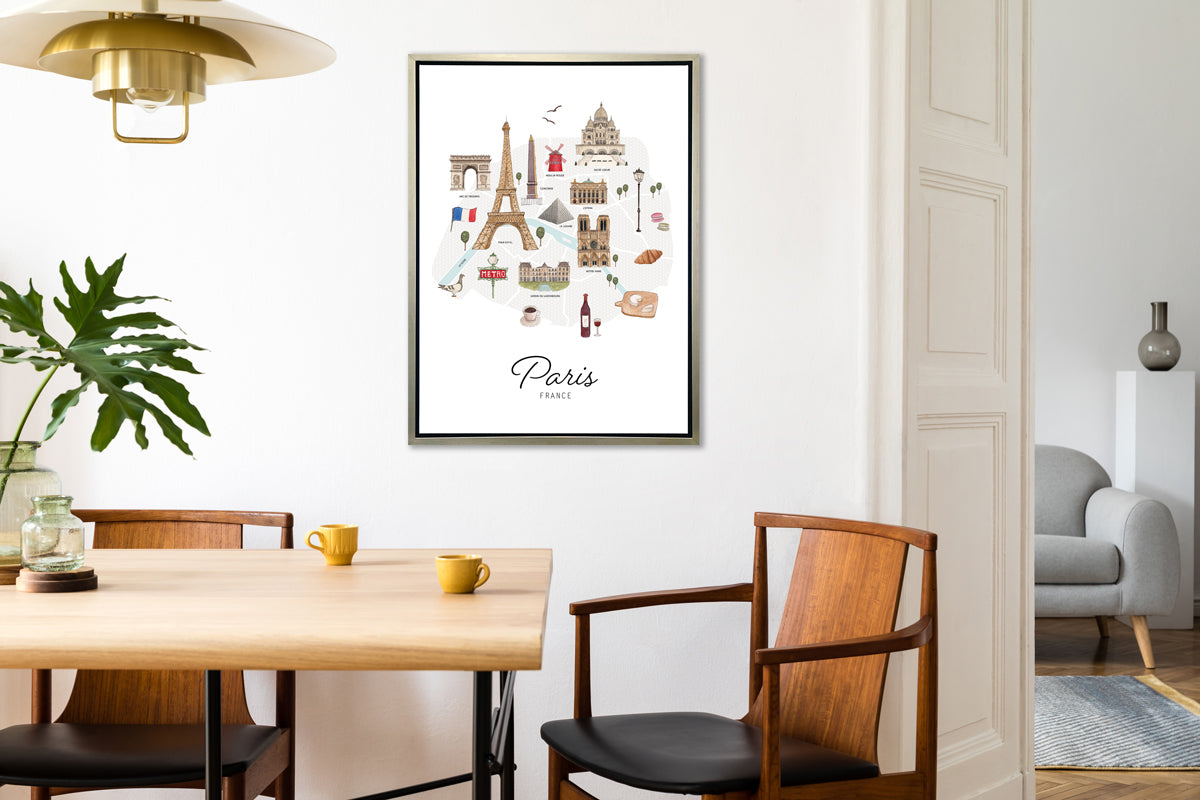 A multicolored map print of Paris, France with the famous landmarks in a silver floater frame hangs on a white wall across from a wood table with two dark yellow coffee mugs, two wood chairs and an opening to another room.