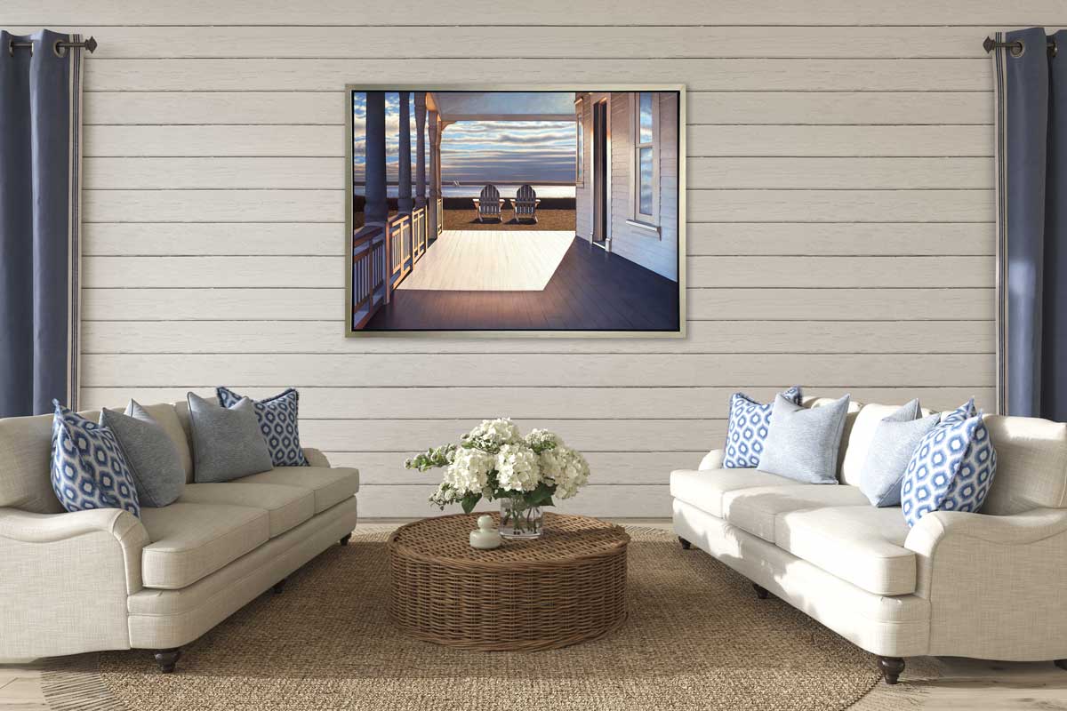 A print of the sunlit porch of a house and two adirondack chairs overlooking the water in a silver floater frame hangs on a white shiplap wall across from two white sofas with blue pillows and a round woven coffee table with white hydrangeas in a clear vessel.