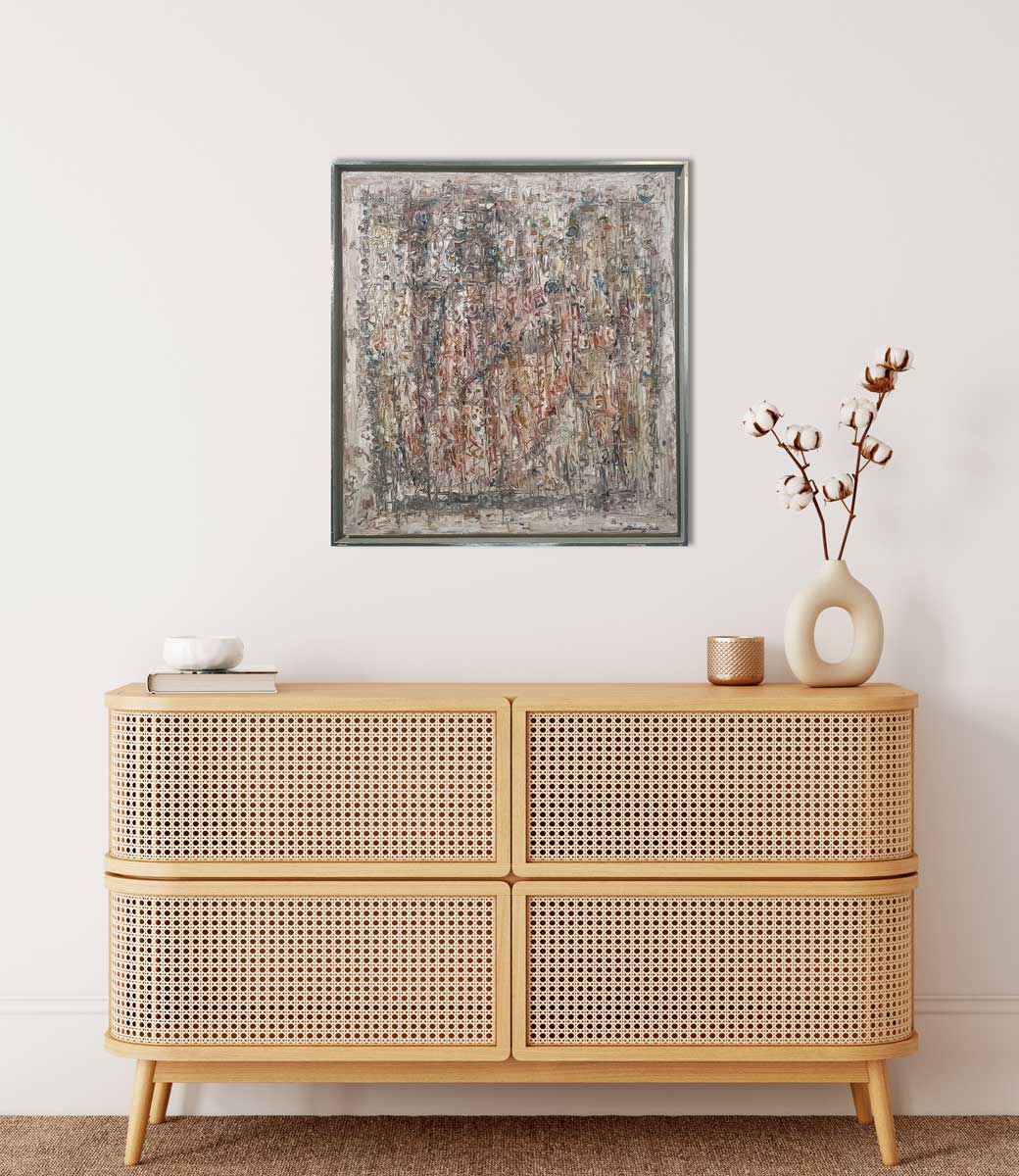 A white abstract painting in a silver floater frame hangs on a white wall above a natural wood console cabinet with a book with a small white bowl, a gold candleholder and a white vase with cotton stems.