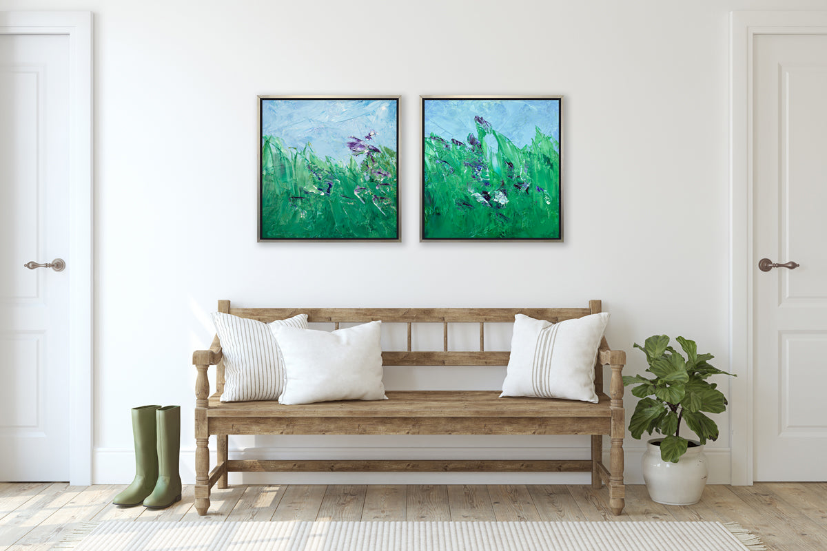 Two blue, green and purple abstract landscape prints in silver floater frames hang side by side on a white wall above a wood bench with a back, armrests and neutral pillows, a small potted plant and a pair of green rubber boots on the floor.