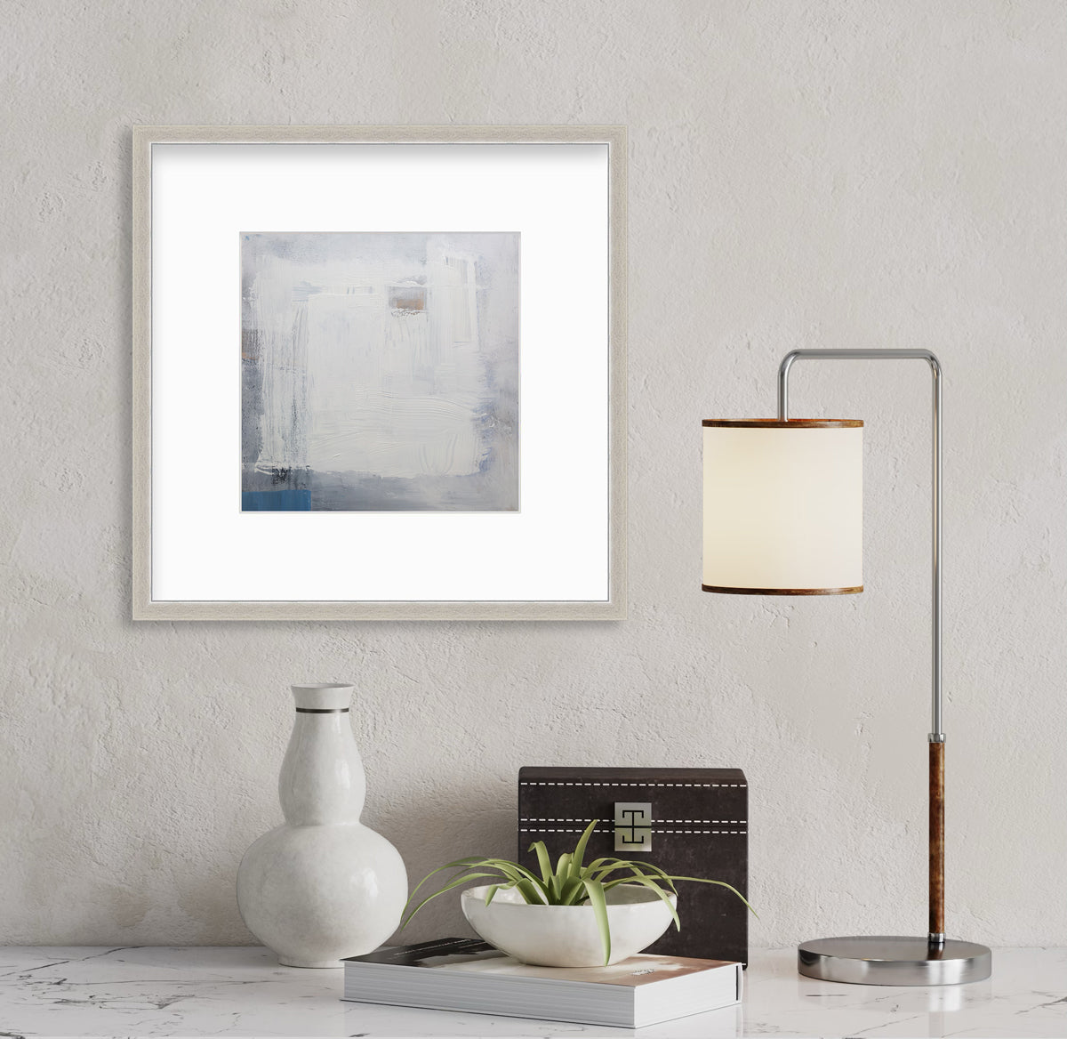 A white, grey and blue abstract print in a silver frame with a mat hangs on a white textured wall above a white marbled surface with a small lamp, a book and some decorative accessories.