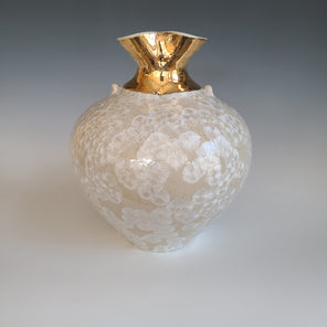A white and cream glazed vessel with two very small handle accents and a fluted gold opening sits on a white surface.