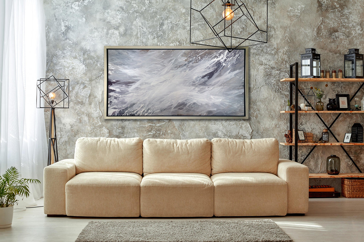 A white, grey and dark grey abstract print in a silver floater frame hangs on a grey textured wall above a beige sofa, modern geometric lighting fixtures and a 4 shelf bookshelf with decorative accessories.