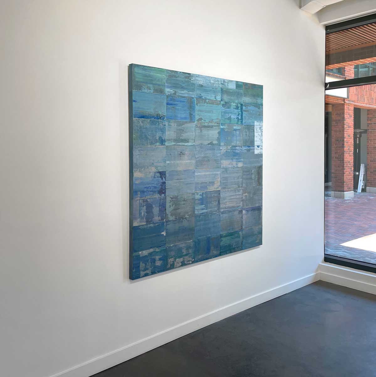 An abstract grid painting with a blue and green wash by Ned Martin hangs on a white wall with a view from the left.