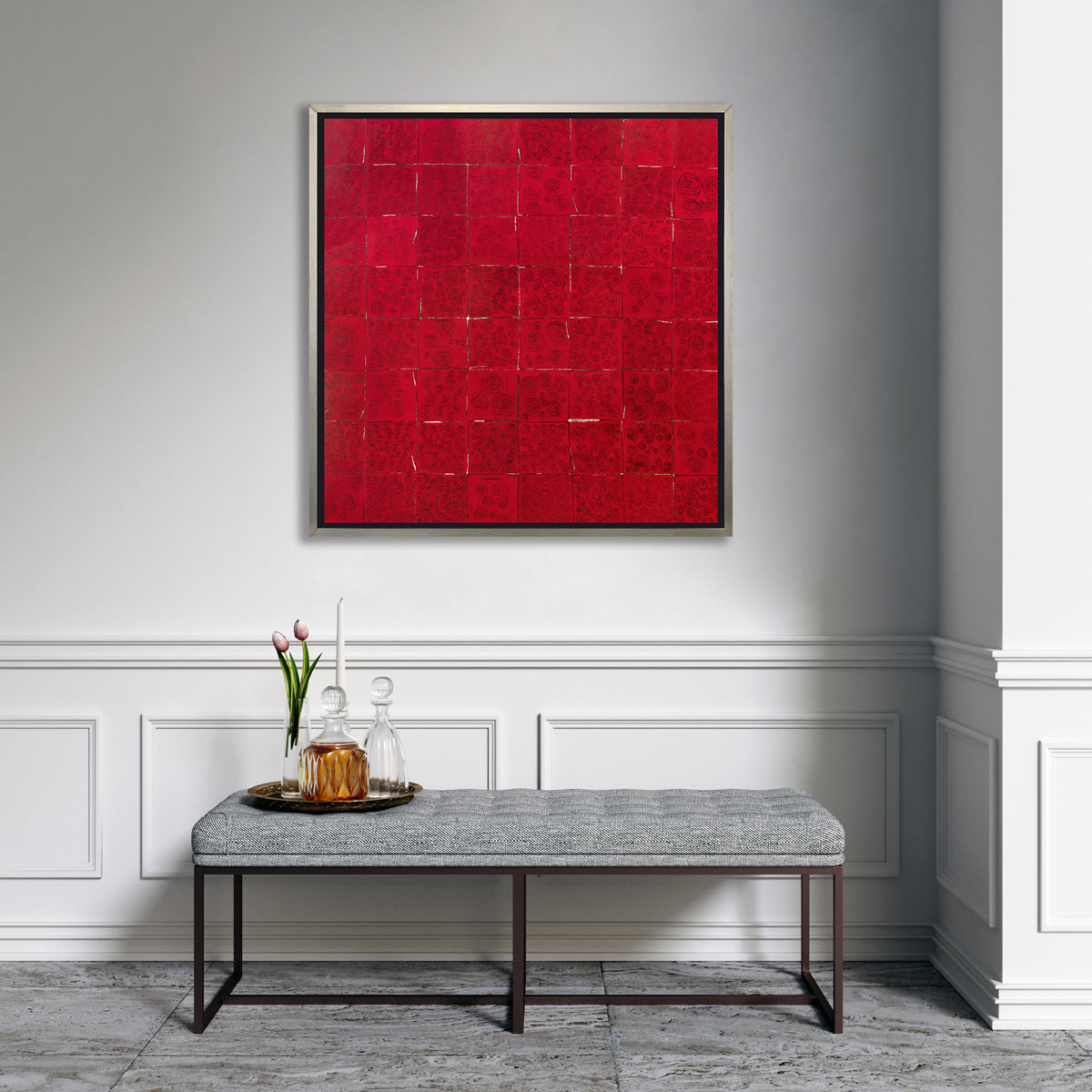 A red and black abstract geometric print in a silver floater frame hangs on a white wall above a grey tufted bench with a tray with clear decanters, a white tapered candle and a clear vase with two pink tulips.