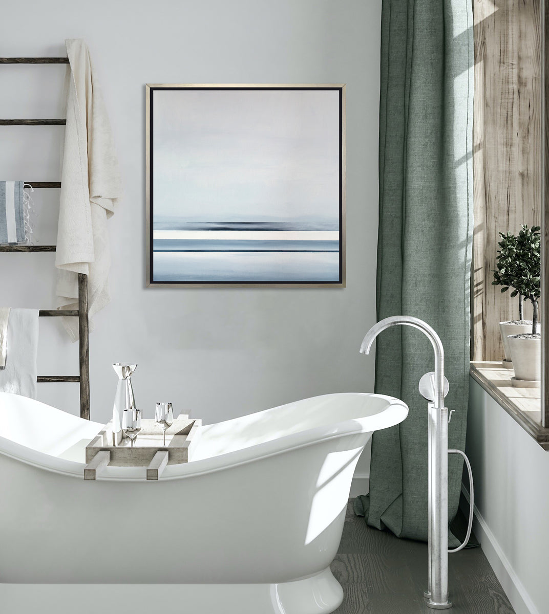 Different sized bands of hues of blue and white hangs in a silver floater frame above a white soaking tub, next to a ladder with towels and a windowsill with two potted plants.