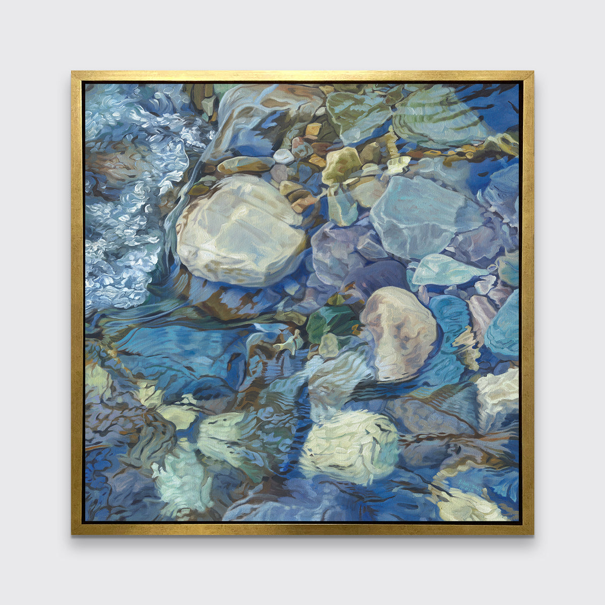 Abstracted print of grey, brown, green and blue river rocks under water hangs in a gold floater frame on a white wall.
