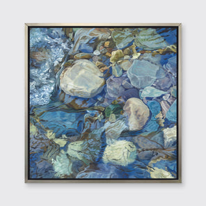 Abstracted print of grey, brown, green and blue river rocks under water hangs in a silver floater frame on a white wall.