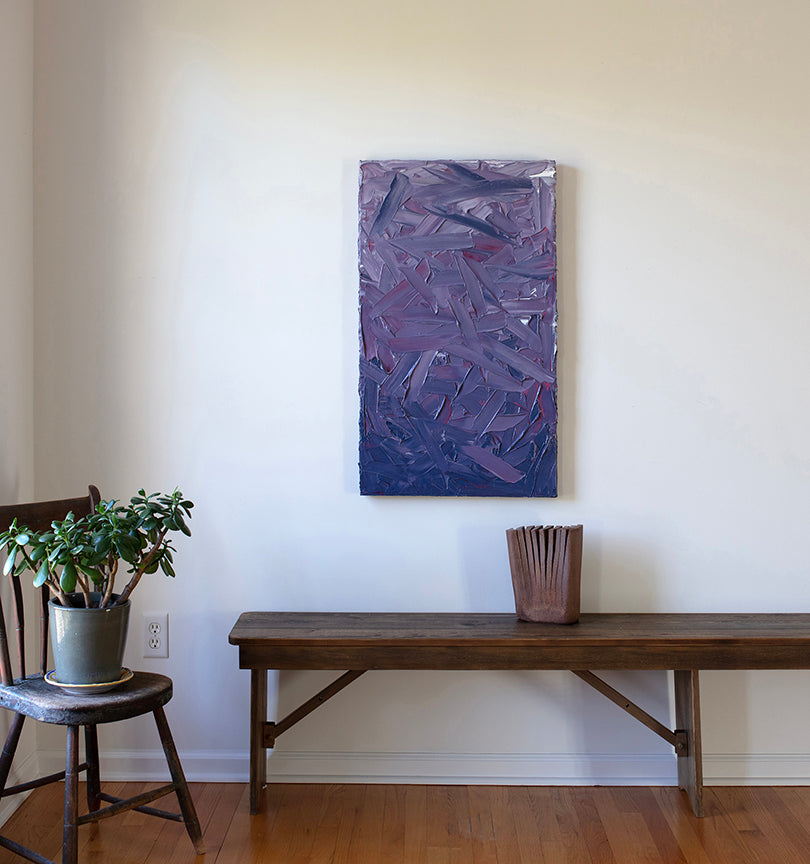 A tonal purple and pink textured abstract painting by Teodora Guererra hangs on a white wall above a wood bench with a wood tabletop sculpture and a chair with a potted plant.