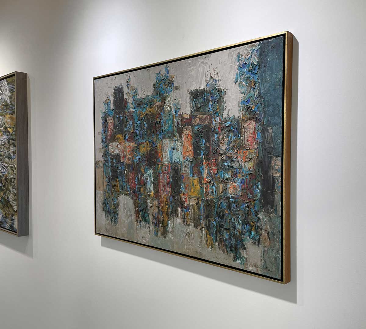 A right side-angle view of a multicolored abstract painting in a warm gold floater frame hangs on a white wall.