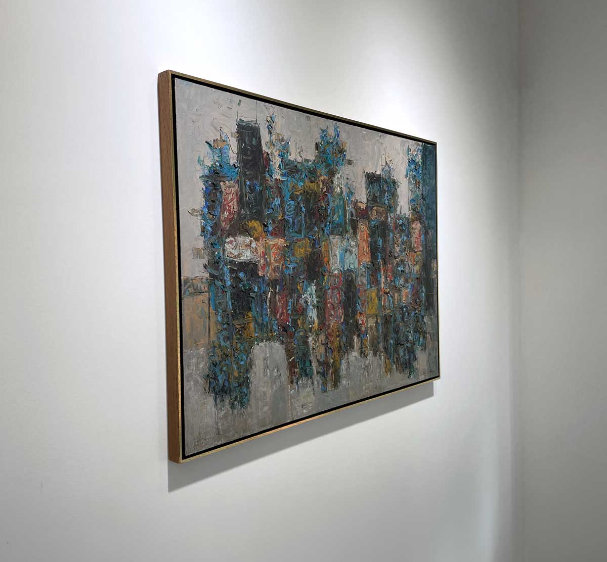 A left side-angle view of a multicolored abstract painting in a warm gold floater frame hangs on a white wall.