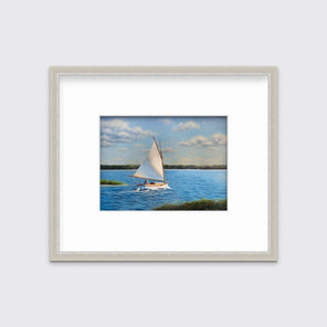 A blue and green realist print of a sailboat on the water surrounded by land masses in a silver frame with a mat hangs on white wall.