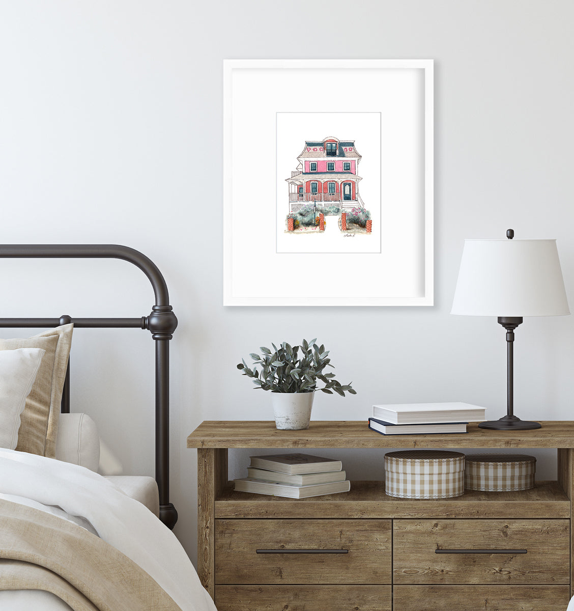 A pink and red house print in a white frame with a mat hangs on a white wall above a wood side table with stacks of books, a small table lamp, a potted plant and next to a bed with a black chrome headboard and neutral bedding.