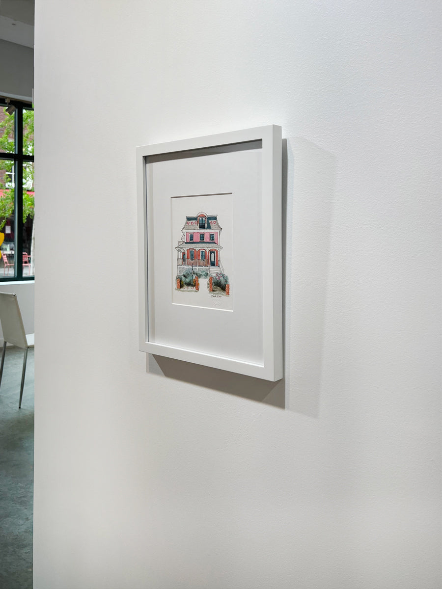 A right side-angle view of a pink, beige, red and slate blue illustration drawing of a house in a white frame hangs on a white wall.