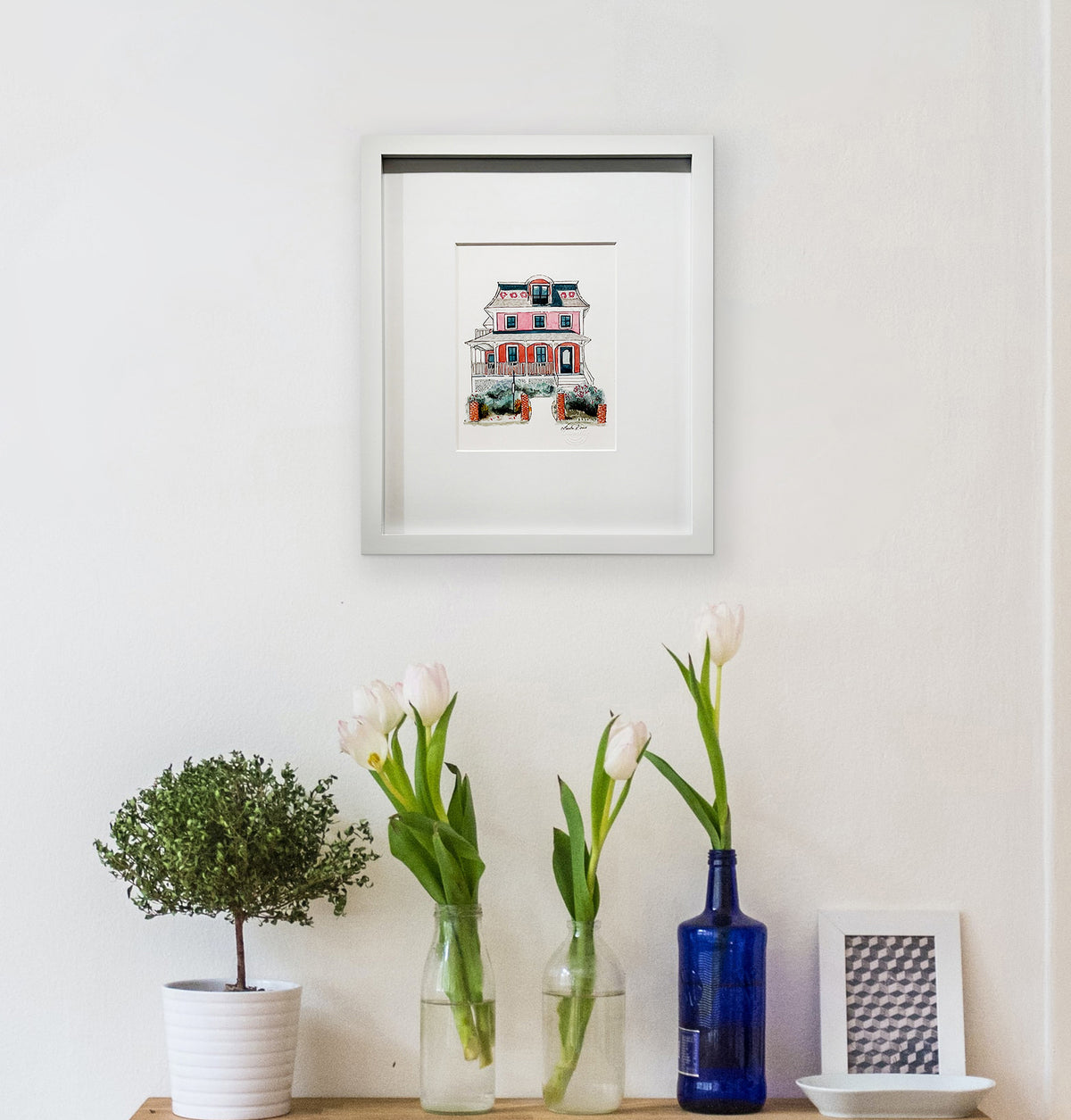 A pink, beige, red and slate blue illustration drawing of a house in a white frame with a mat hangs on a white wall above a wood surface with a few bottled white tulips, a potted shrub and a white dish.