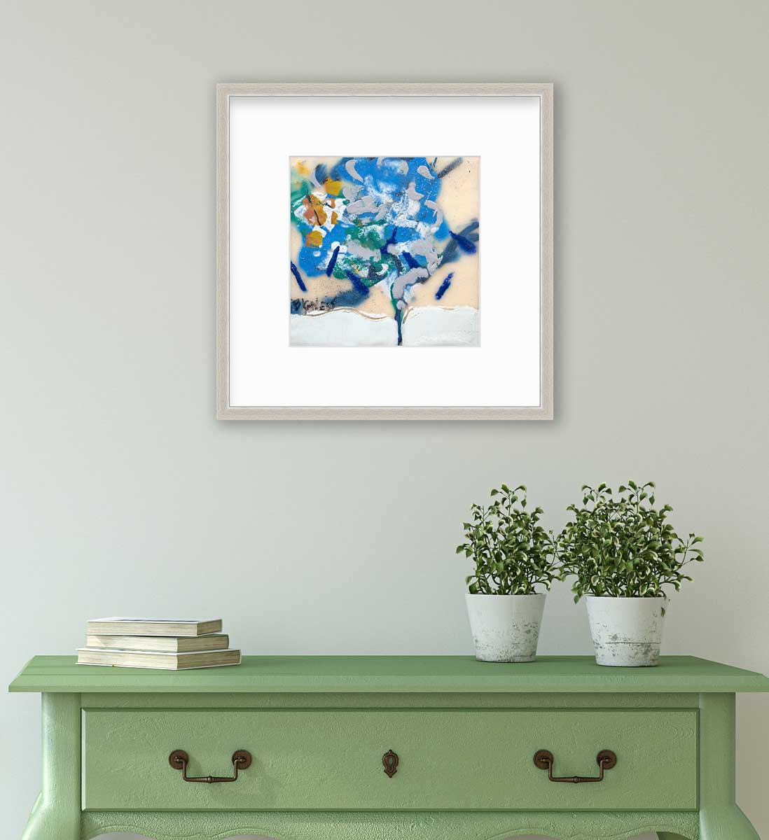 A blue, white, green and orange abstract print in a silver frame with a mat hangs on a white wall above a green console table with a stack of books and two potted plants.