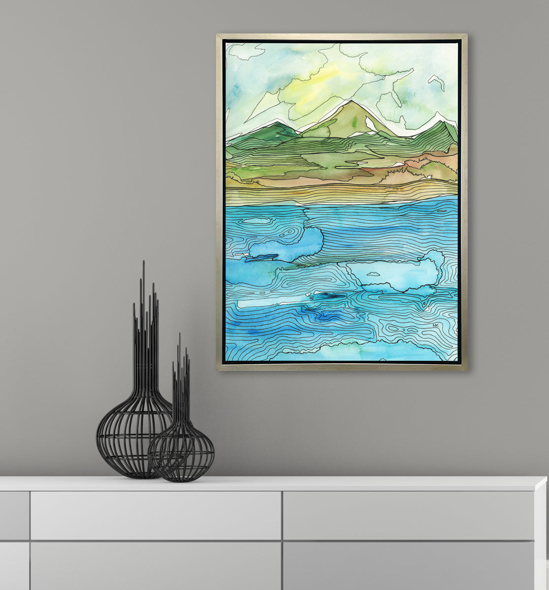 A blue, green, brown and light yellow abstract landscape with black outlines print in a silver floater frame hangs on a grey wall above a white cabinet with two black modern decorative accessories.