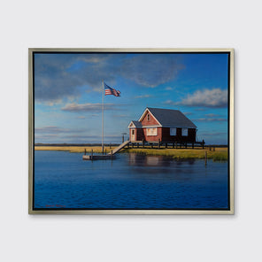 A print of a red bay house looking over a body of water in a silver floater frame hangs on a white wall.