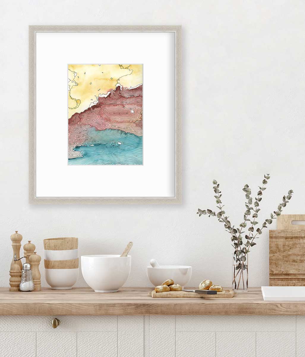 A yellow, dark red, teal and black abstract landscape print in a silver frame with a mat hangs on a white wall above a butcher block countertop with bowls, a cutting board and a clear glass vase with eucalyptus stems.