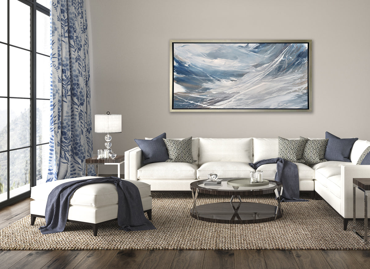 A grey, white and blue abstract print in a silver floater frame hangs on a white wall above a white sectional sofa with blue pillows, a round glass coffee table and a oversized matching white ottoman with a blue throw blanket.