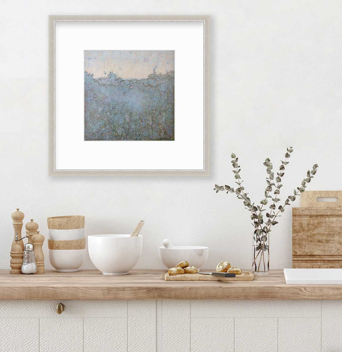 A dark abstract landscape print in a silver frame with a mat hangs on a white wall above a butcher block countertop with bowls, a cutting board and clear glass vase with eucalyptus stems.