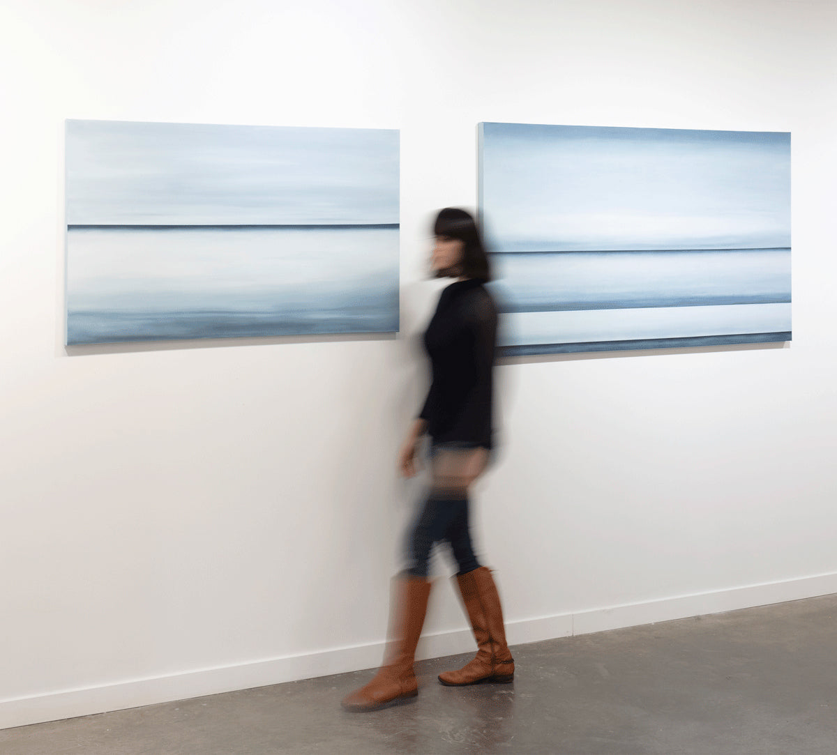 A woman in a dark outfit walks in front of two abstract blue paintings hanging on a white wall.