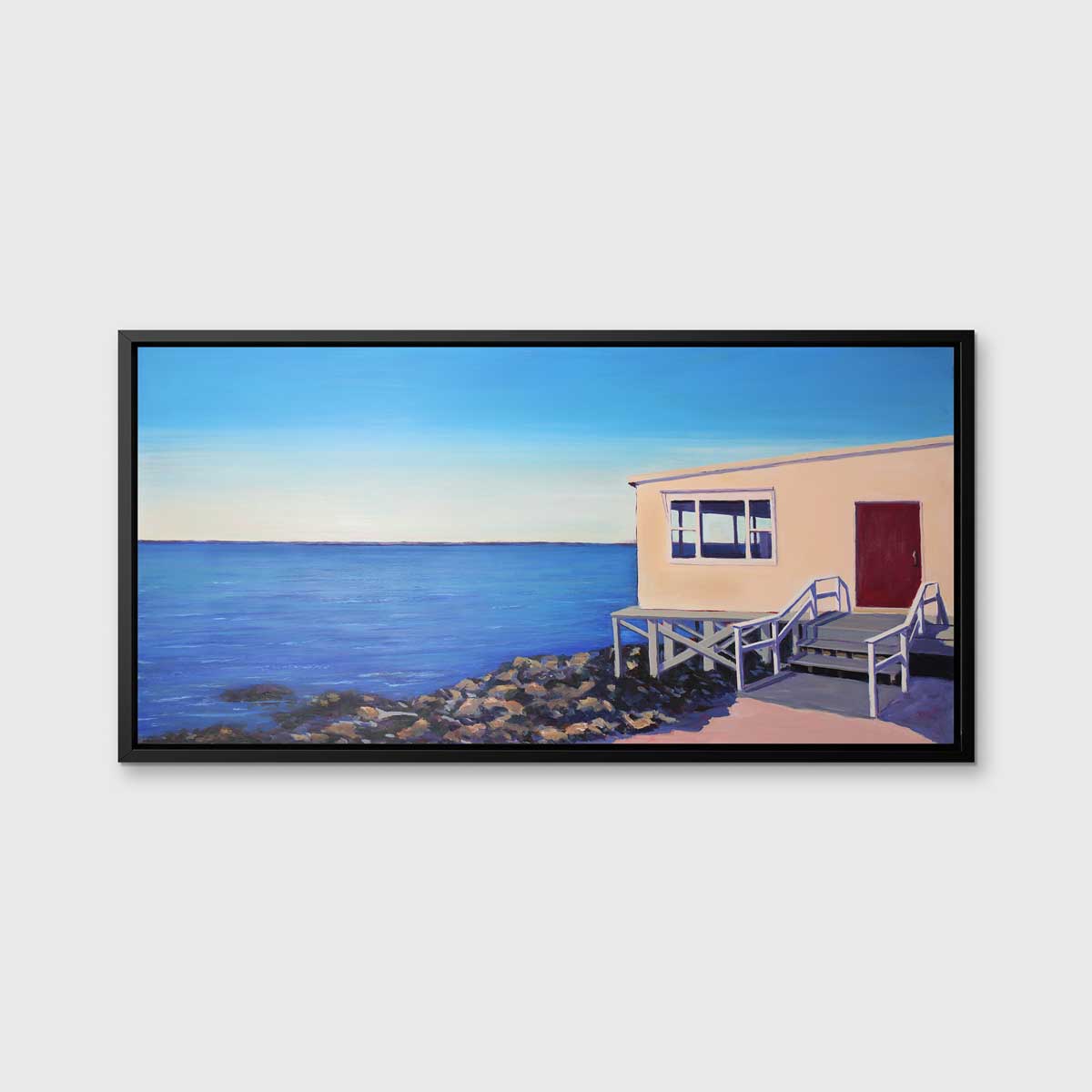 A blue, beige and grey contemporary seaside print with a beach house on the rocks next to the ocean in a black floater frame hangs on a white wall.