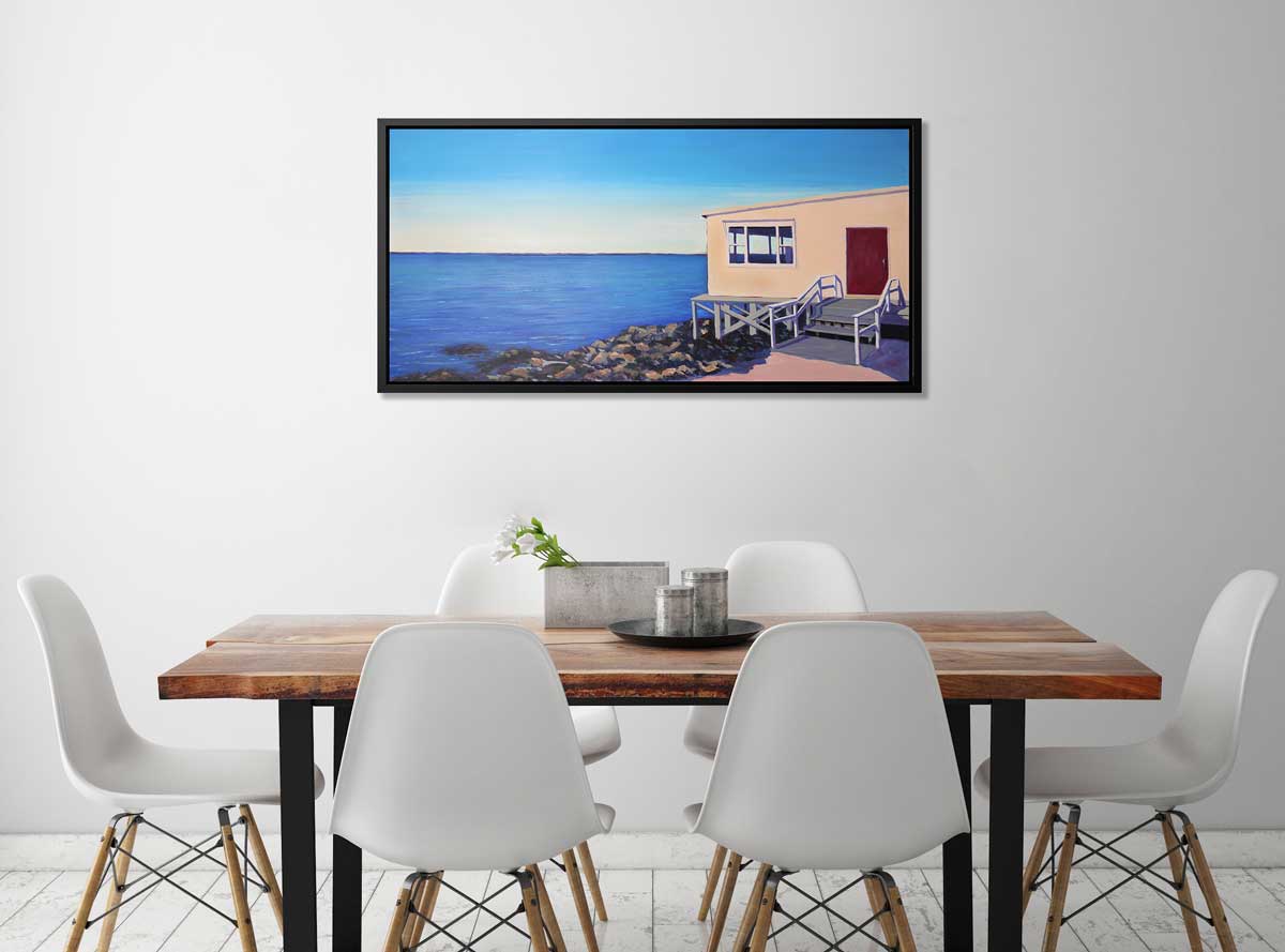 A blue, beige and grey contemporary seaside print with a beach house on the rocks next to the ocean in a black floater frame hangs on a white wall above a live edge table with six white chairs surrounding.