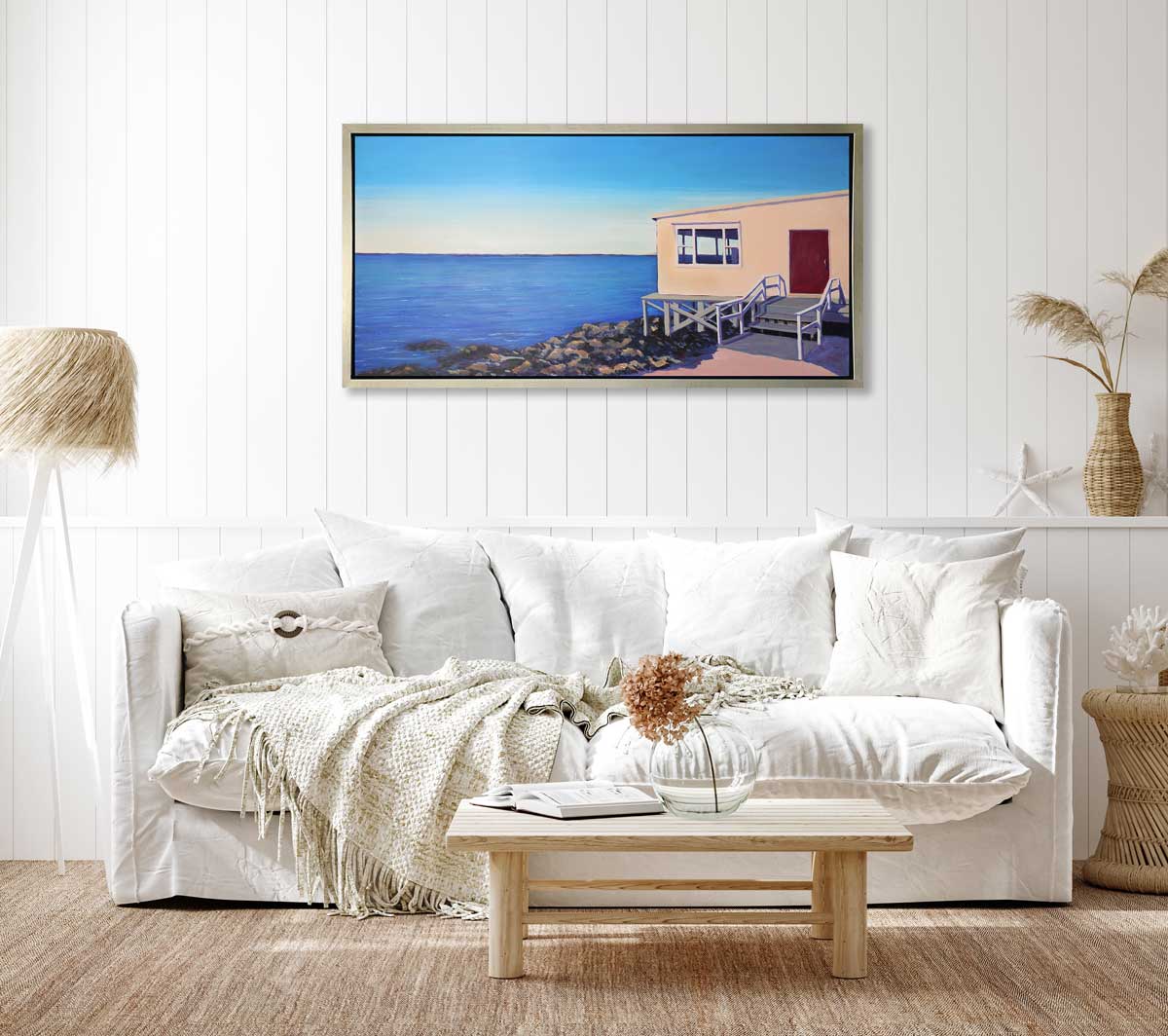 A blue, beige and grey contemporary seaside print with a beach house on the rocks next to the ocean in a silver floater frame hangs on a white wall above a white sofa with pillows, a small natural wood coffee table and a tripod beach grass floor lamp.