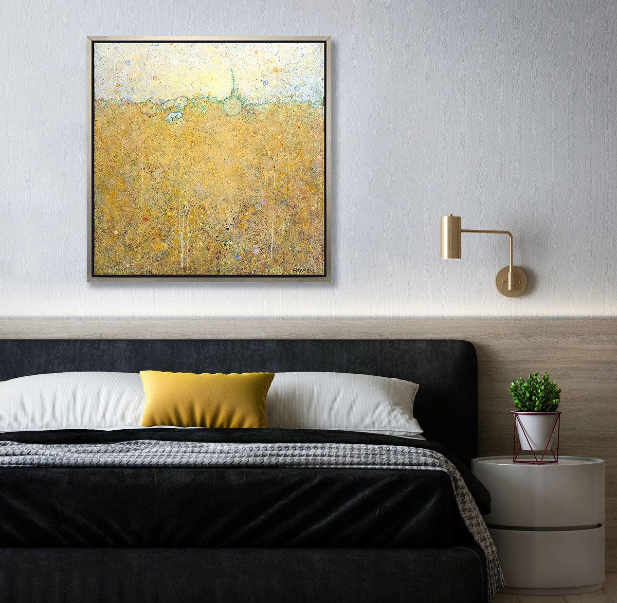 A dark yellow abstract landscape print in a silver floater frame hangs on white wall above a black headboard, black bedding with a yellow pillow and a round white side table with a potted plant.