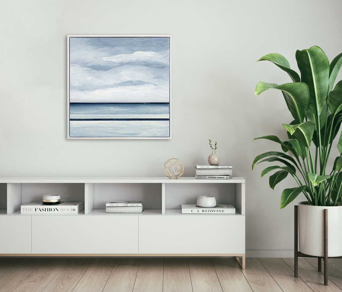 A blue and white abstract landscape print in a white floater frame hangs on a white wall above a modern white credenza with several stacked books, some small decorative accessories and a potted floor plant.