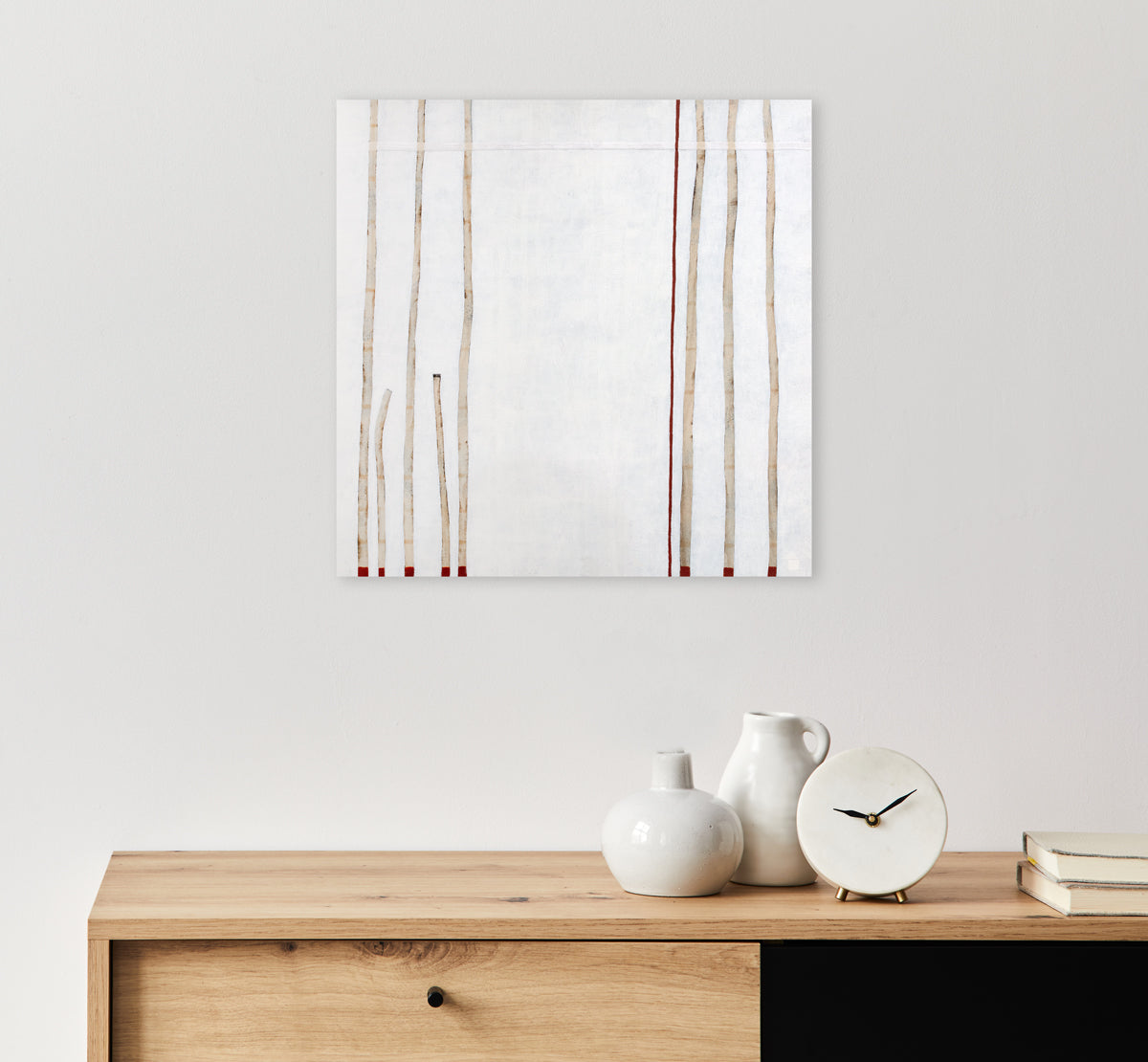 A minimalist painting of beige lines on the right and left of the composition with hints of red on a white background hangs on a white wall above a natural wood console with white ceramic vases and a clock sitting on top.