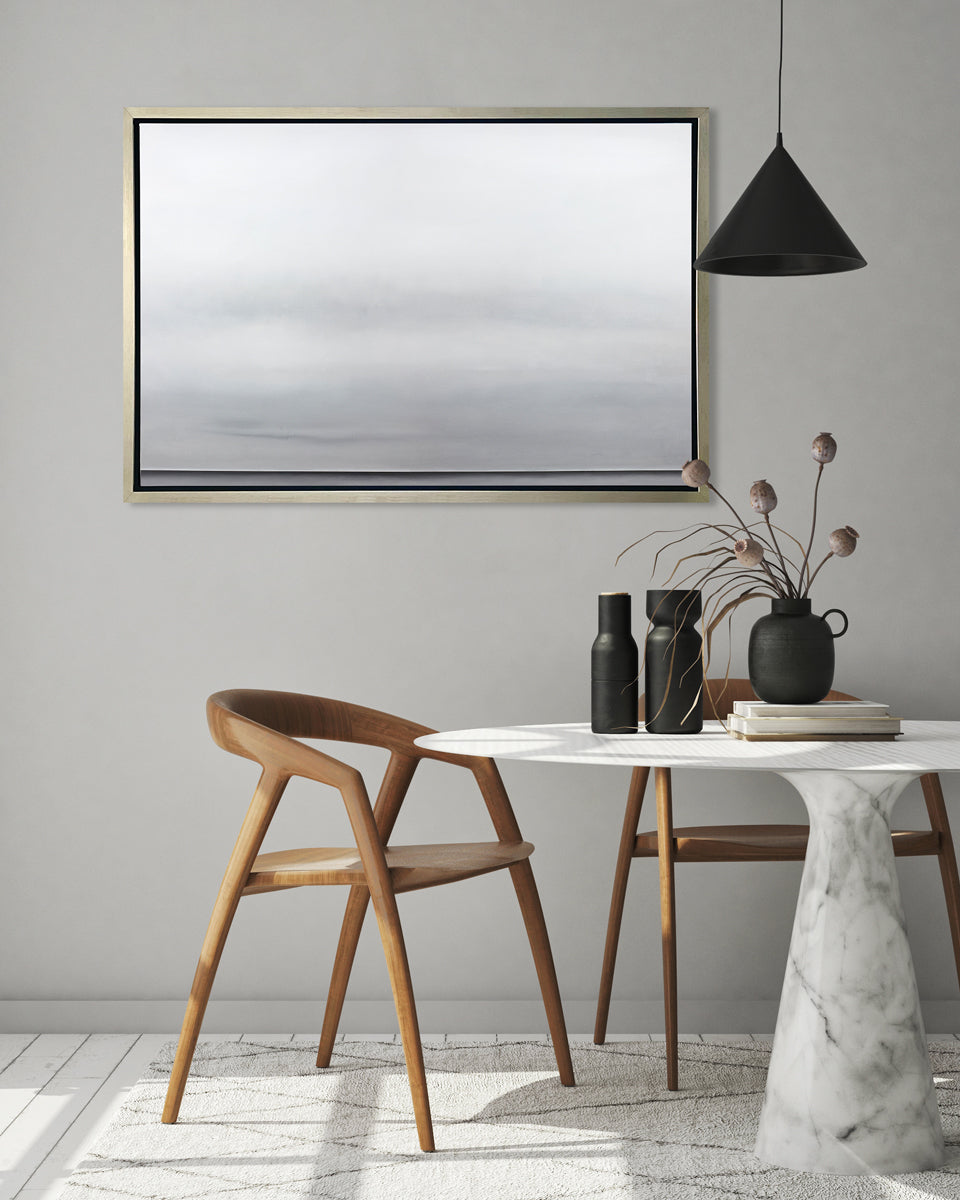 A tonal grey abstract landscape print in a silver floater frame hangs on a grey wall above a round white marble pedestal table with black vases and two modern wood chairs.