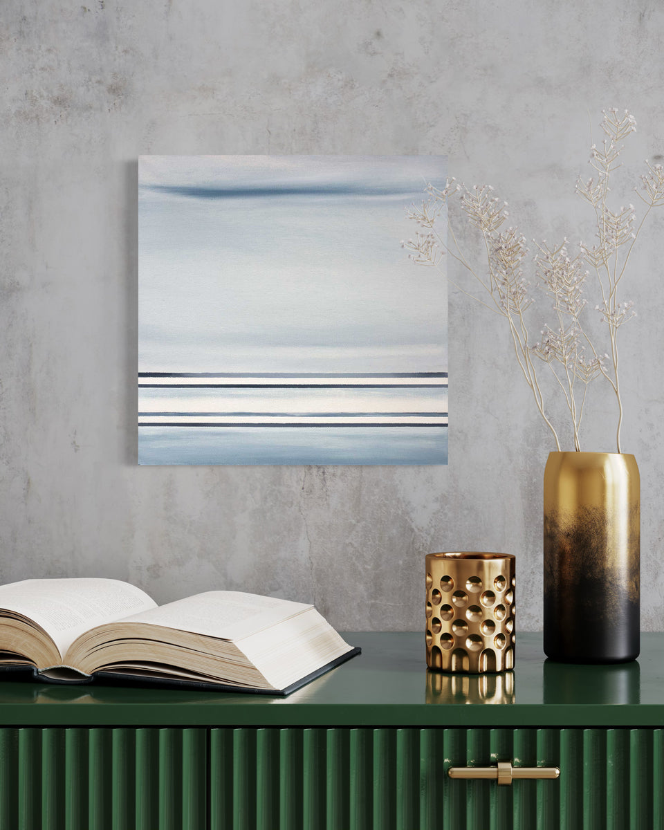 A blue and white linear abstract painting hangs on a grey textured wall above a green credenza with an open book, a gold candleholder and a gold and black vase with white floral stems.
