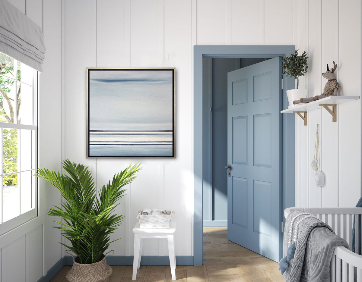A blue and white linear abstract print in a silver floater frame hangs above a large floor plant and a side table with baby accessories and to the right is a blue door and a crib.