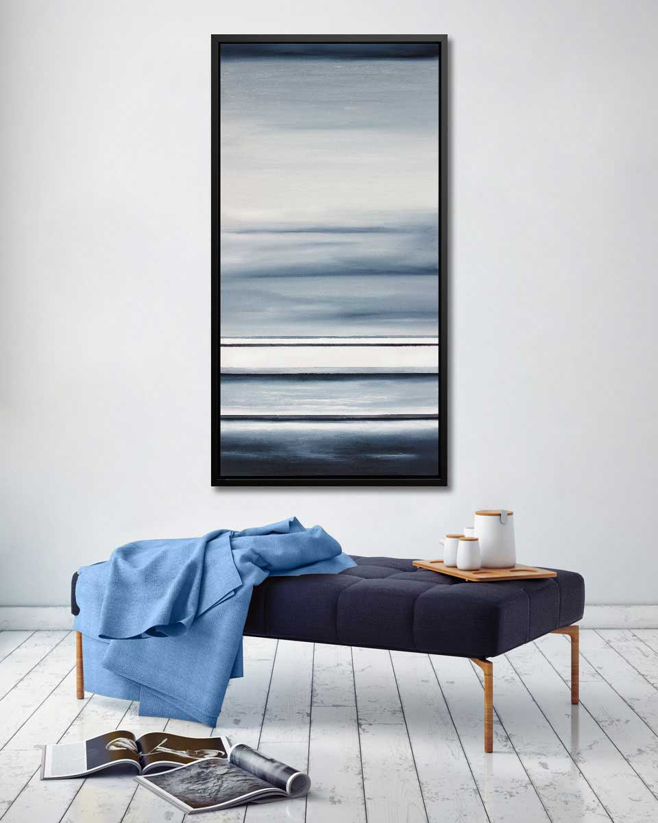 Different sized bands of hues of blue and white hanging in a black floater frame above a dark blue tufted bench with a blanket draped over and a tea set on a small wooden tray.
