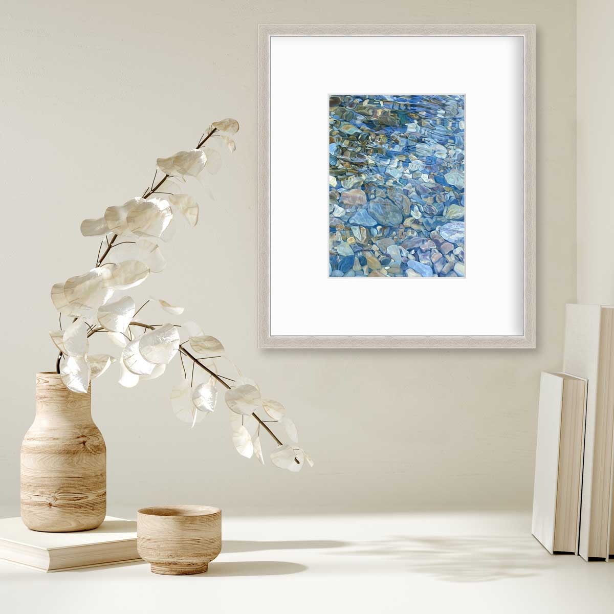 Abstracted river rocks under water hangs in a silver frame with a mat on a white wall above a stack of books and a wooden vase with white leaf stems.