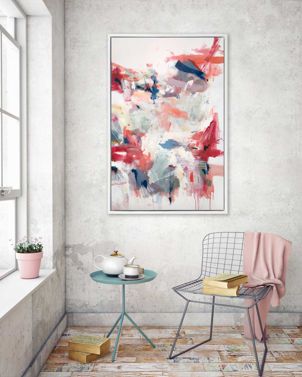 A pink, blue and white abstract print in a white floater frame hangs on a grey textured wall above a grey wire chair, a small teal side table and stacks of books.