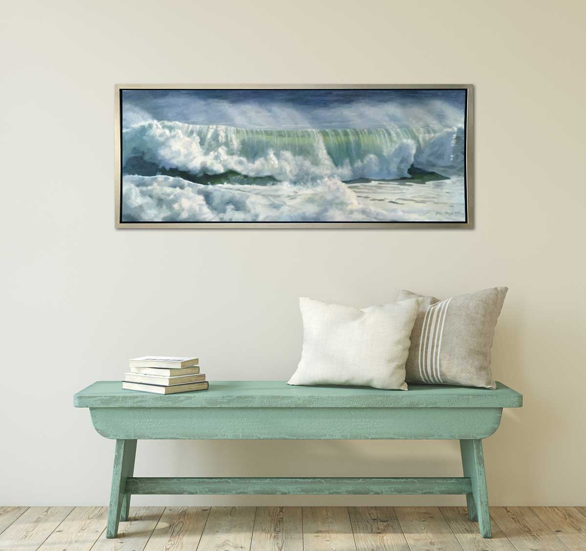 A blue, white and green abstract rolling wave coming to shore print in a silver floater frame hangs on a white wall above a seafoam green bench with two neutral pillows and a stack of books.
