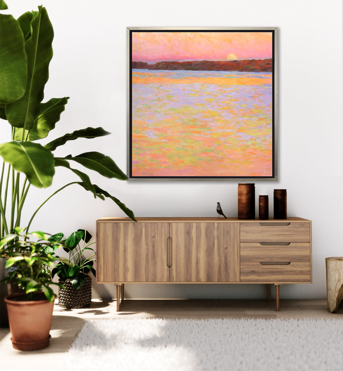 A pink, orange, lavender and red impressionist landscape print in a silver floater frame hangs on a white wall above a wood credenza with three dark wood cylindrical vases, a small bird figurine and several potted floor plants.